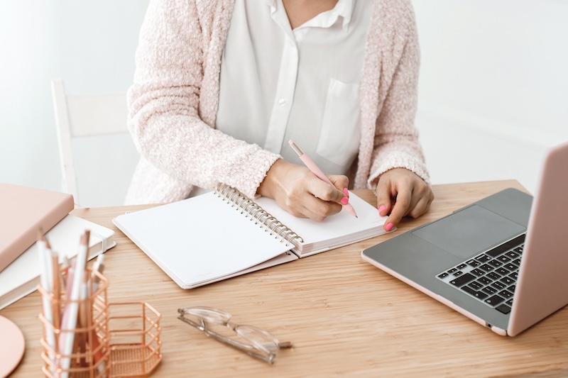 woman writing in a notepad on desk with laptop in shades of pink