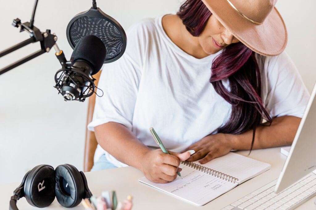 woman sitting at desk writing in a notebook with laptop and microphone