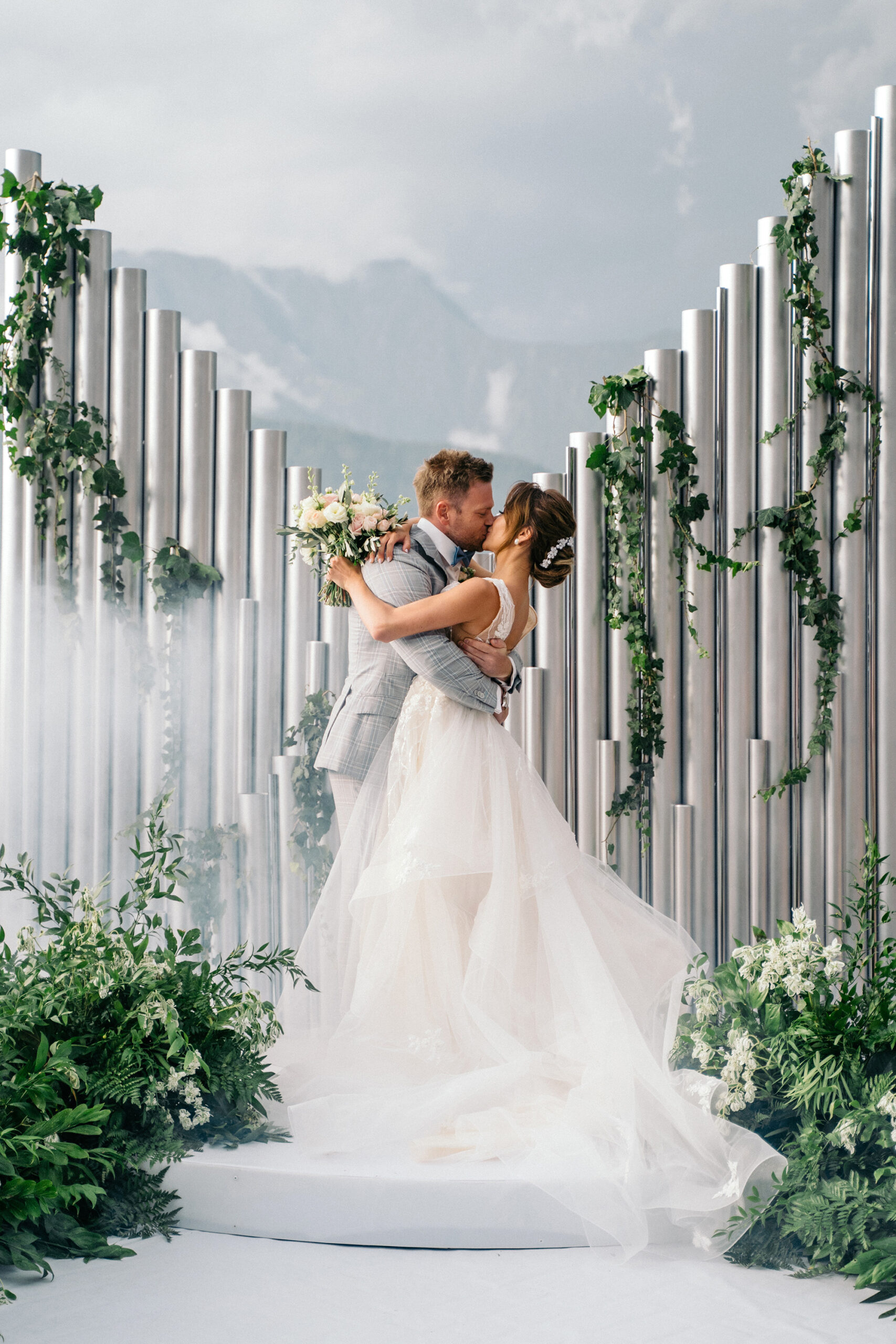 a bride and groom kissing during their luxury outdoor wedding ceremony, surrounded by greenery in front of some silver columns
