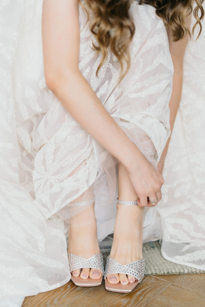 Bride in beaded white wedding dress putting on her silver beaded wedding heels